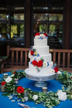 a white wedding cake with red, white and blue flowers on the top is surrounded by greenery