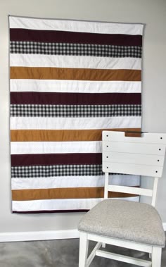 a white chair sitting next to a wall hanging on the side of a wall with a brown and black striped quilt