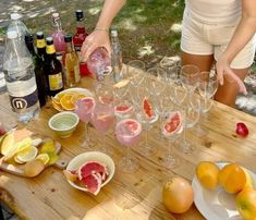 a woman is pouring wine into glasses on a wooden table with fruit and bottles in the background