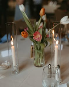 flowers in vases and candles on a white table cloth with silverware at the end