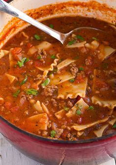 a red pot filled with pasta and meat soup on top of a white wooden table