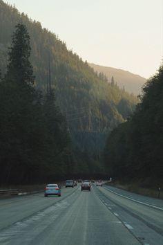 cars are driving down the road in front of some trees and mountains on a sunny day