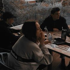 three people sitting at a wooden table with drinks in front of them and one person talking on the phone