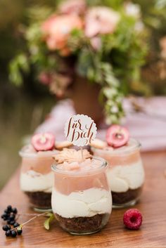 three desserts in small glass jars on a wooden table with flowers and greenery