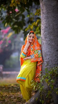 a woman in an orange and yellow outfit leaning against a tree with her hand on her head