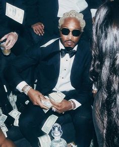 a man in a tuxedo and sunglasses sitting on a couch surrounded by money