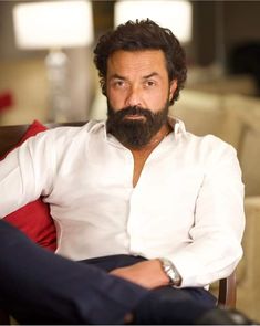 a man with a beard sitting in a chair looking off to the side while wearing a white shirt and blue pants