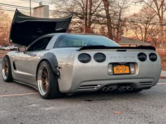 the rear end of a silver sports car parked in a parking lot with its hood open