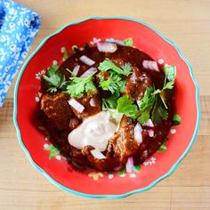 a red bowl filled with meat and vegetables on top of a wooden table next to a blue napkin