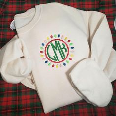 a white sweater with a monogrammed christmas wreath on the front, and a red plaid blanket behind it