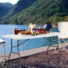 an outdoor table with food and drinks on it near the water in front of mountains