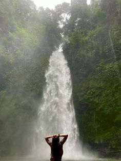 a woman standing in front of a waterfall with her hands on her head and arms behind her head