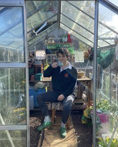 a young man sitting on a chair in a greenhouse drinking from a cup while looking at the camera