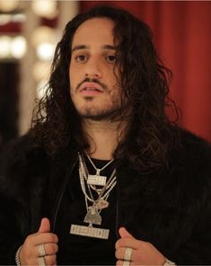 a man with long curly hair wearing a black jacket and holding his hands up in front of him