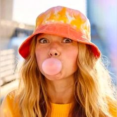 a woman with long blonde hair wearing a red hat and blowing a bubble in front of her face