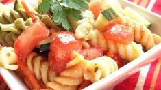 a white bowl filled with pasta and vegetables on top of a red table cloth next to a green leafy garnish