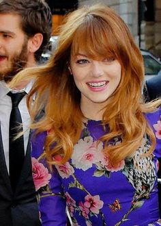 a woman with red hair is smiling and wearing a purple floral dress while standing next to a man in a black suit