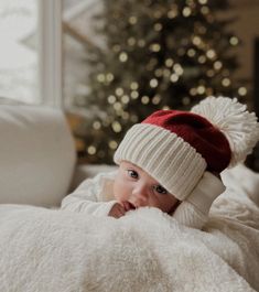 a baby wearing a red and white hat laying on top of a bed next to a christmas tree