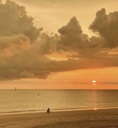 two people are sitting on the beach as the sun sets in the distance behind them