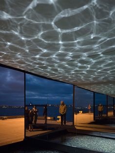 people are standing in front of large windows looking out at the water and buildings with lights on them