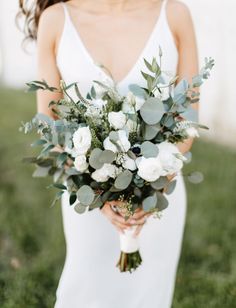 a woman holding a bouquet of flowers in her hands and wearing a white dress with greenery