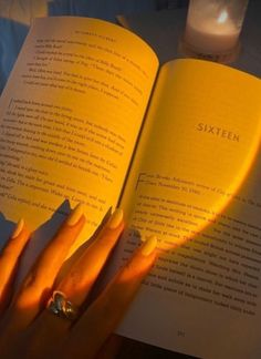 a woman's hand holding an open book with yellow nails and ring on it