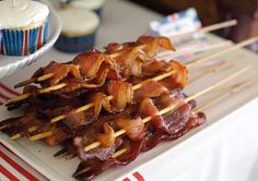 bacon skewers on a plate with cupcakes and other food items in the background