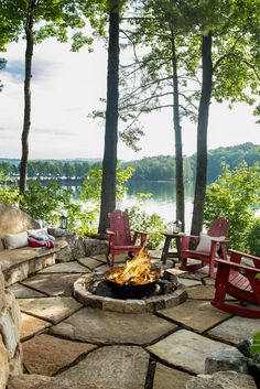 an outdoor fire pit with chairs around it and water in the background, surrounded by trees