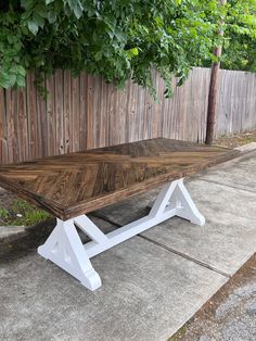 a wooden table sitting on top of cement ground next to a fence and tree with green leaves