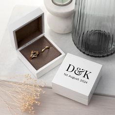 a white box with a gold ring in it next to a vase and some dried flowers