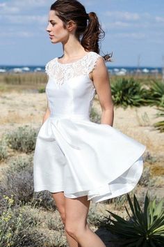 a woman in a short white dress is walking on the sand near some plants and bushes