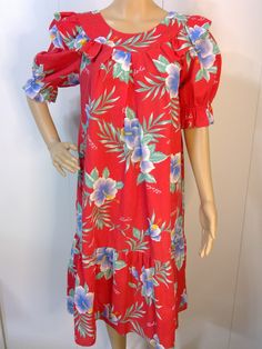 Beautiful Red with Hibiscus Flowers and Aloha Across Fabric.  Ruffle around neckline with criss-cross back.  Short puff sleeves with elastic and ruffle. One pocket. Big 12" ruffle at bottom. 65% Cotton/35% Poly  VGVC with no flaws noted. Size M - Refer to Measurements: Bust/38" Waist/56" Hip/Free Length/40" Smoke & Pet Free Studio Red Hawaiian Dress For Spring, Red Hibiscus Print Dress For Vacation, Red Hawaiian Floral Print Dress, Hawaiian Style Red Floral Print Dress, Red Hawaiian Dress With Floral Print, Red Hawaiian Beach Dress, Tropical Short Sleeve Dress For Holiday, Red Tropical Short Sleeve Dress, Aloha Dress