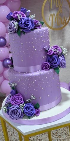 a three tiered cake with purple flowers on top and balloons in the back ground