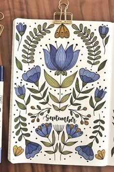 an open notebook with blue flowers on it next to some markers and pens, along with a clipboard