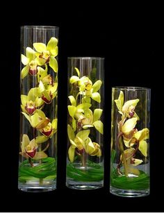 three glass vases with yellow flowers and green grass in them on a black background