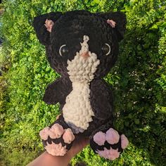 a hand holding a black and white crocheted teddy bear in front of green bushes