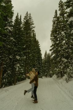 two people walking down a snow covered road in the middle of pine trees and evergreens