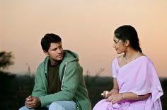 a man sitting next to a woman on top of a grass covered field at sunset