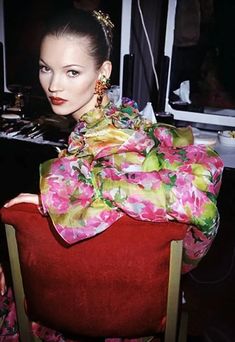 a woman sitting on top of a red chair in front of a pink wall and wearing a flowered scarf