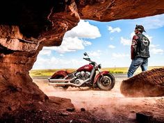 a man standing next to a red motorcycle in the middle of a dirt road under a rock formation