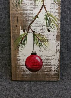 a red ornament hanging from a pine tree branch on a wooden plaque with white paint