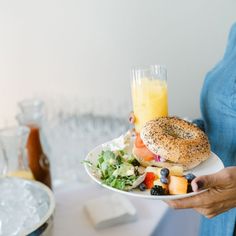 a woman holding a plate with a bagel and fruit salad on it, next to a glass of orange juice