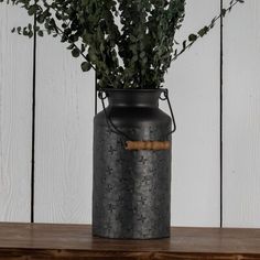 a metal vase filled with green plants on top of a wooden table next to a white wall