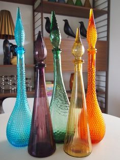 four different colored glass vases sitting on top of a white table in front of a shelf