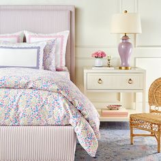 a bedroom with pink and white bedding, wicker chair, lamp and dresser