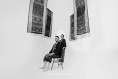 two men sitting on chairs in front of some rugs hanging from the ceiling above them