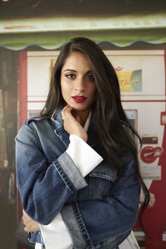 a woman wearing a jean jacket standing in front of a vending machine with her hand on her chin