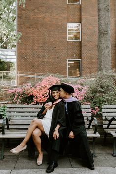 a man and woman in graduation gowns sitting on a park bench with flowers behind them