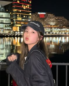 a woman standing in front of a body of water at night wearing a black jacket and hat