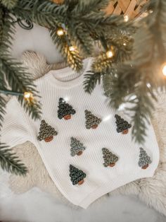 a white sweater with christmas trees on it sitting next to a pine tree and some lights